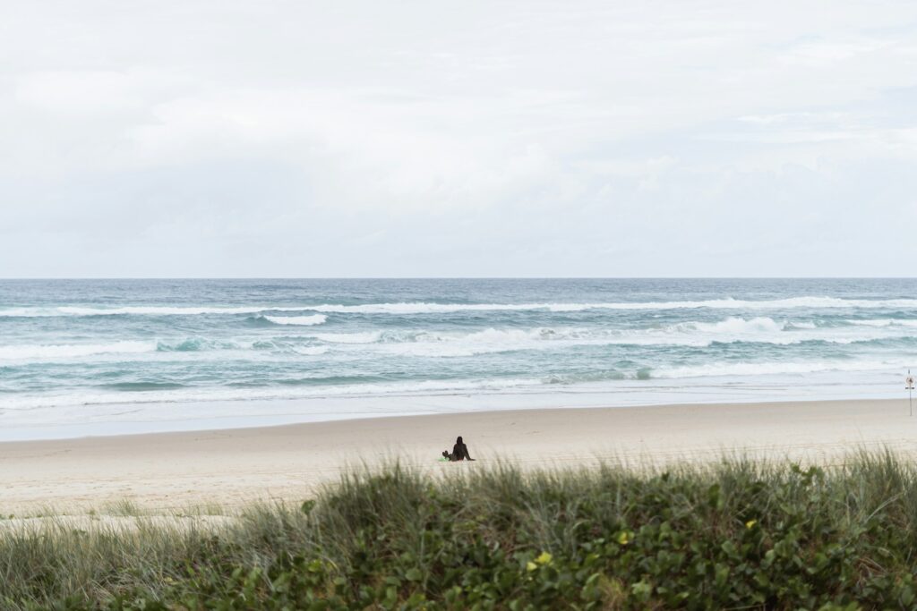 A solitary figure sitting on the beach, waves gently rolling in (symbolizing the “slow waves of loss”).
